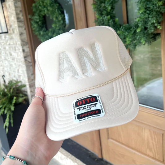 A hand holds a cream-colored trucker cap with large embroidered 'AN' lettering on the front. The cap features a structured curved brim, mesh back panels, and a prominent 'OTTO TRUCKER' patch on the side.